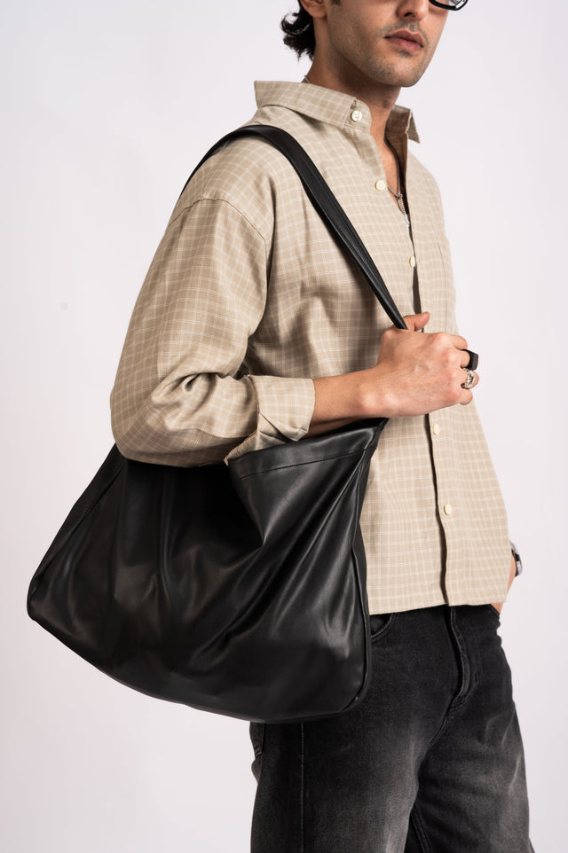 A person wears a relaxed-fit beige Nomad Check Boxy Shirt- Beige and black jeans while carrying a large black slouchy shoulder bag.