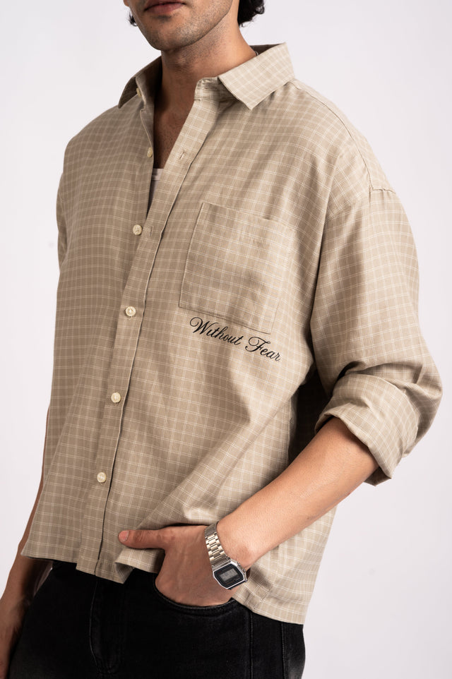 A person wearing a Nomad Check Boxy Shirt- Beige, a relaxed-fit shirt with a windowpane check pattern and the words Without Fear embroidered on the chest pocket.